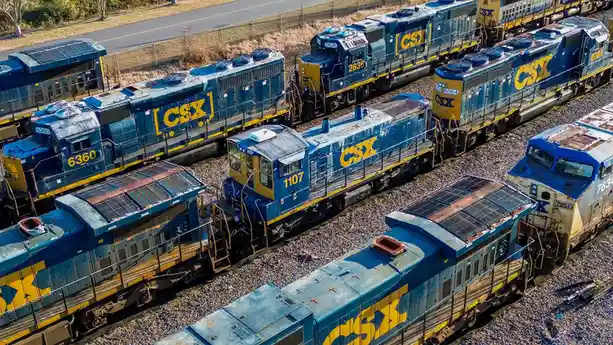 An aerial photo shows multiple blue and yellow CSX locomotives parked on parallel tracks in a rail yard, some showing signs…