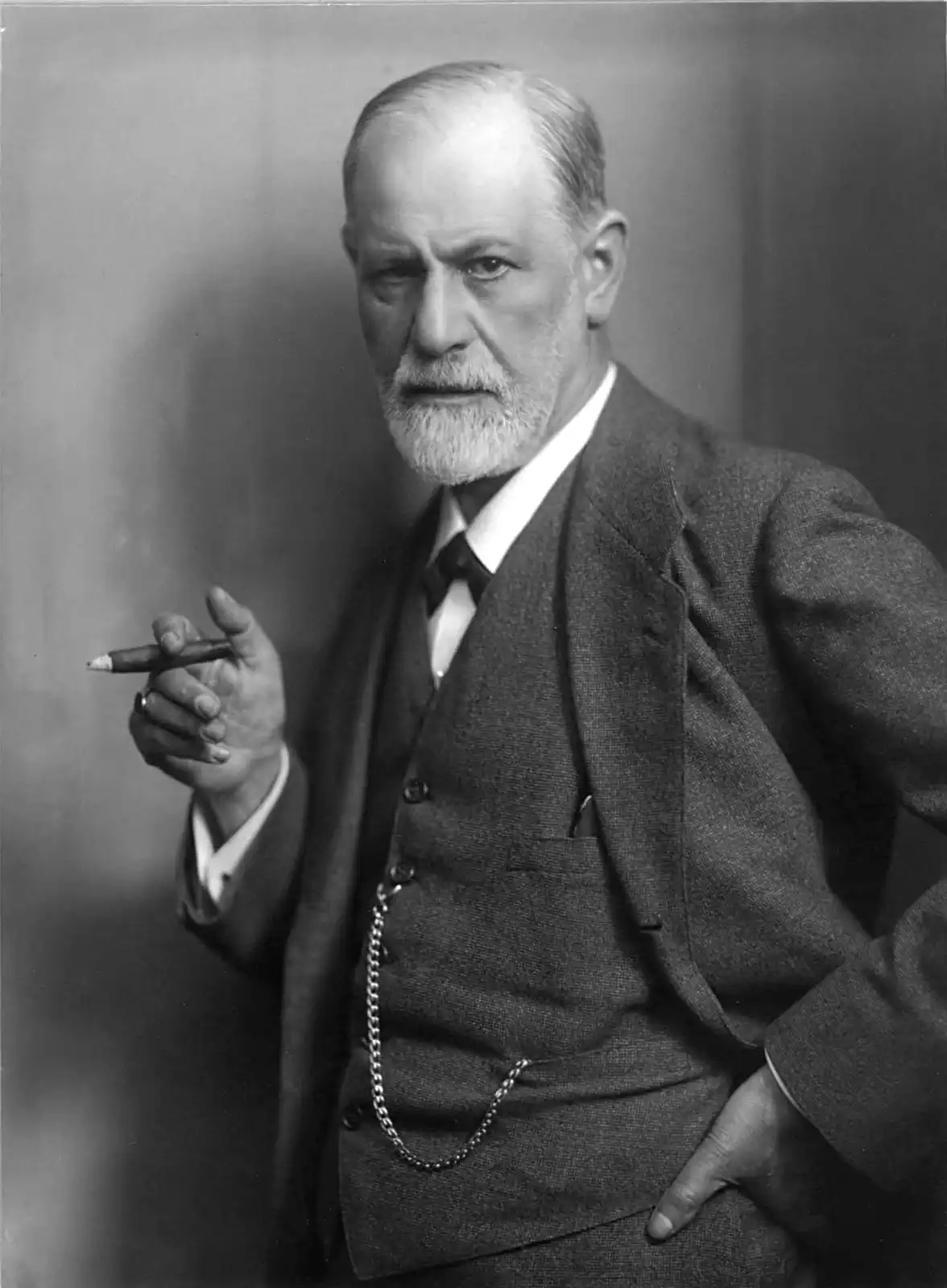 a black-and-white portrait of Sigmund Freud, an older man with a white beard, wearing a suit and vest, holding a cigar in…