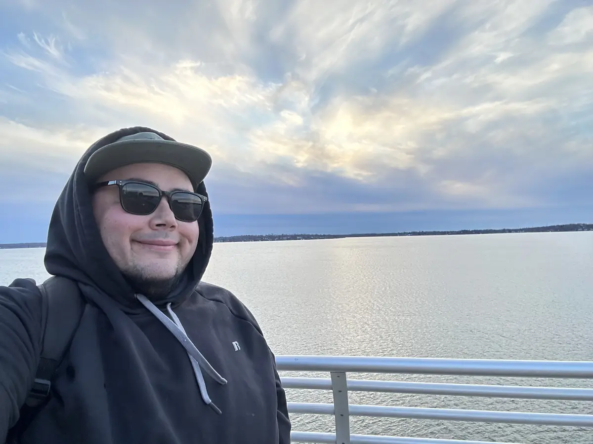 A man wearing a dark hoodie with the hood up, a baseball cap, and sunglasses, takes a selfie on a railing overlooking a…
