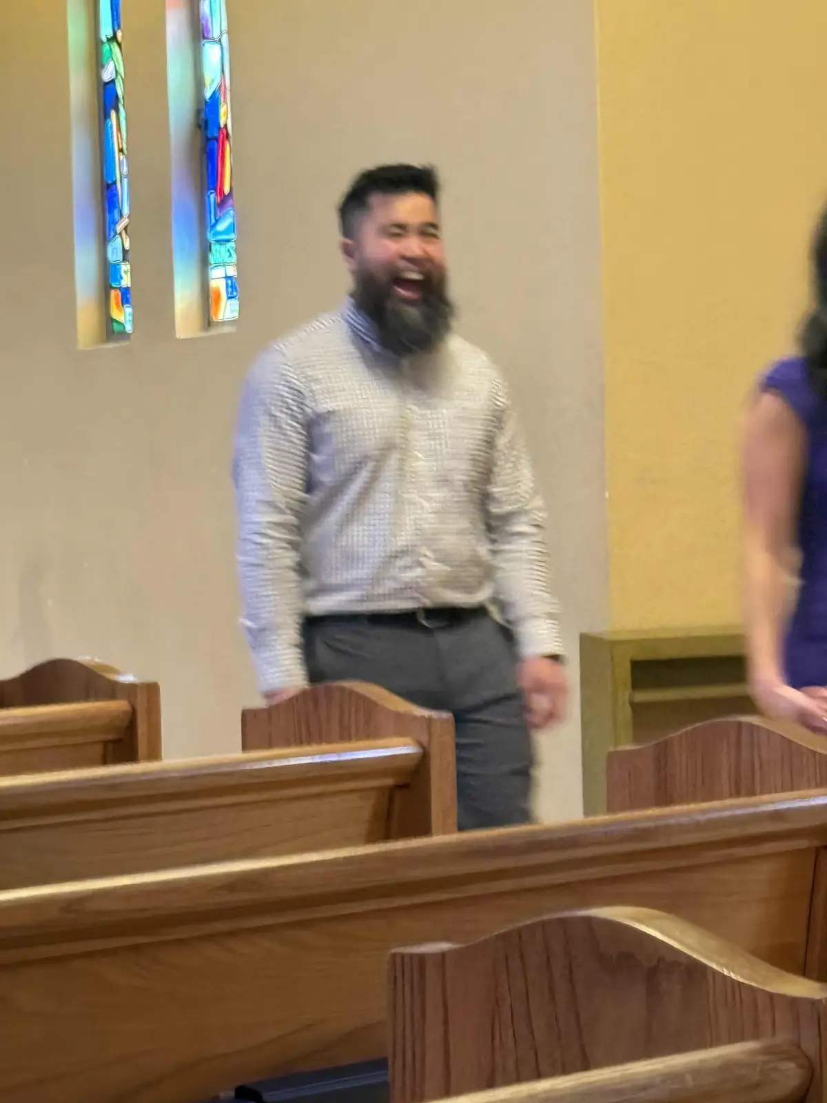 A blurry photo shows a bearded man in a church pew laughing heartily, with stained glass windows visible behind him. The…