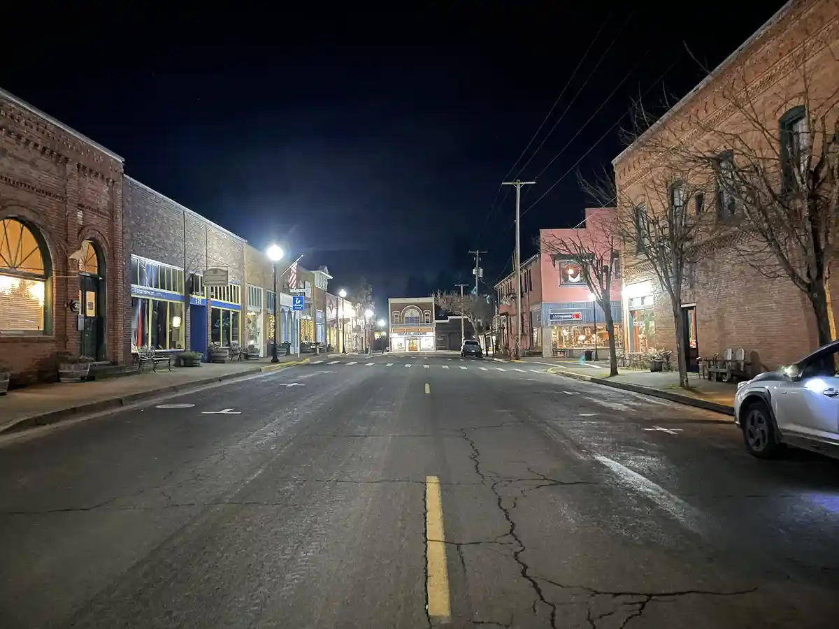 A nighttime photo of a quiet, empty main street in a small town, lined with brick buildings and storefronts, with a single…