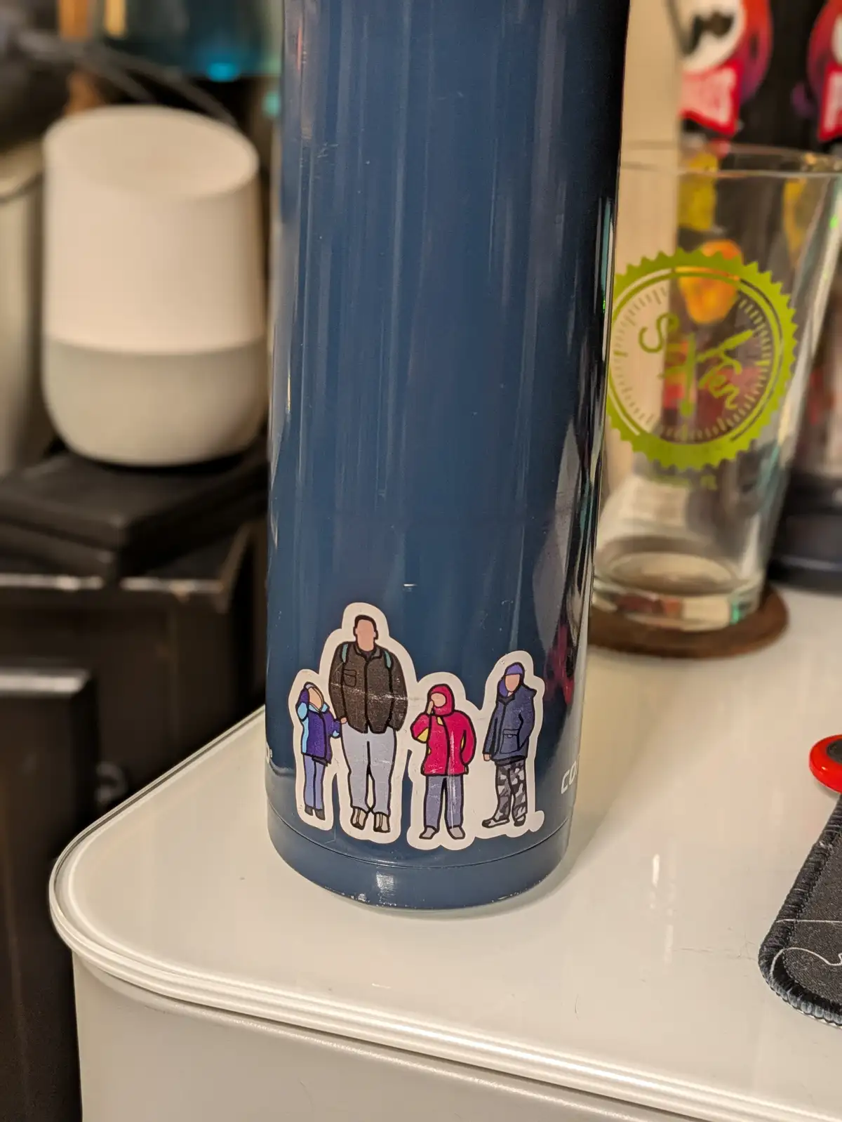 A blue insulated thermos sits on a white countertop, decorated with a sticker of four cartoon figures: an adult male holding…