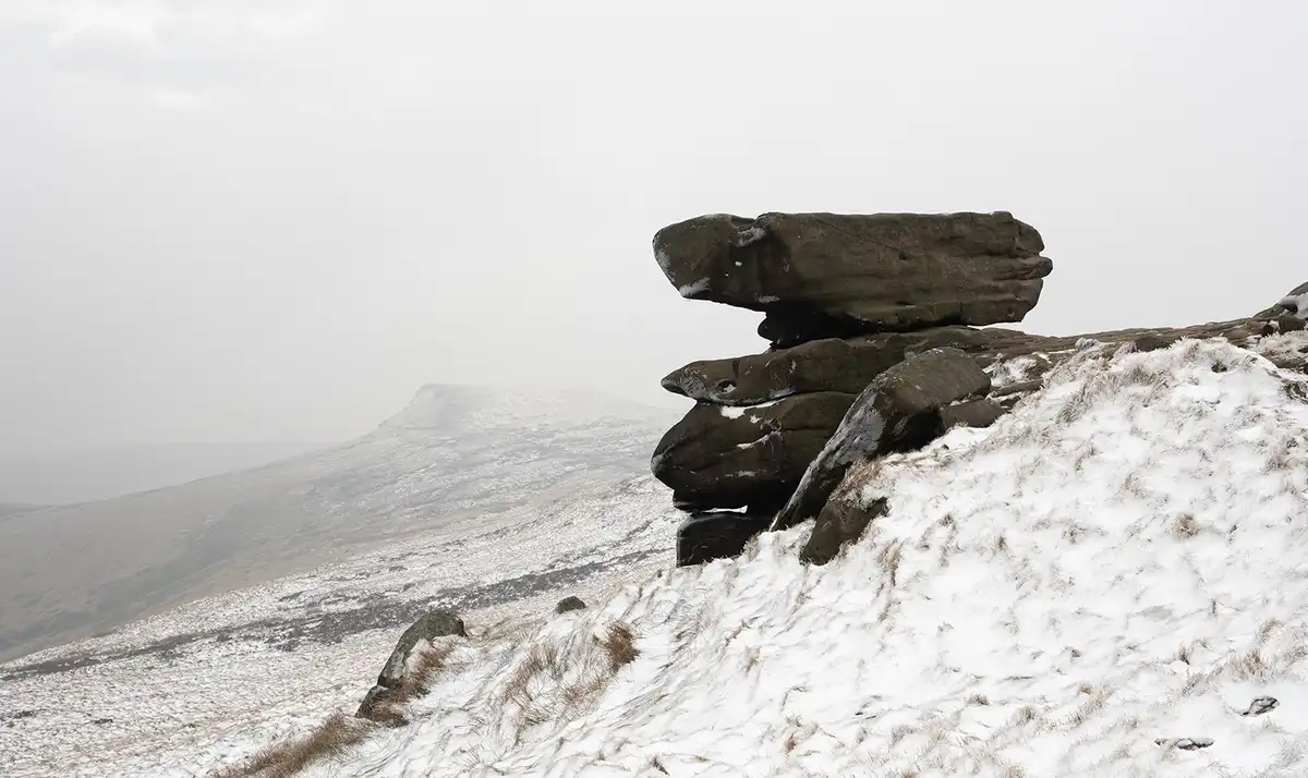A snowy, misty mountain landscape features a prominent, precarious stack of large, dark rocks balanced on a snow-covered…