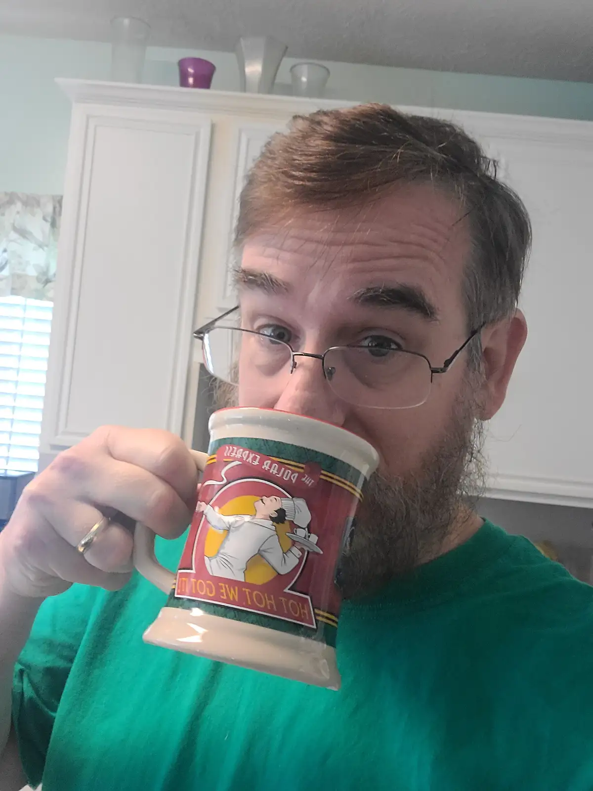 A man with glasses and a beard holds a mug up to his mouth, obscuring most of his face. The mug features a cartoon chef and…