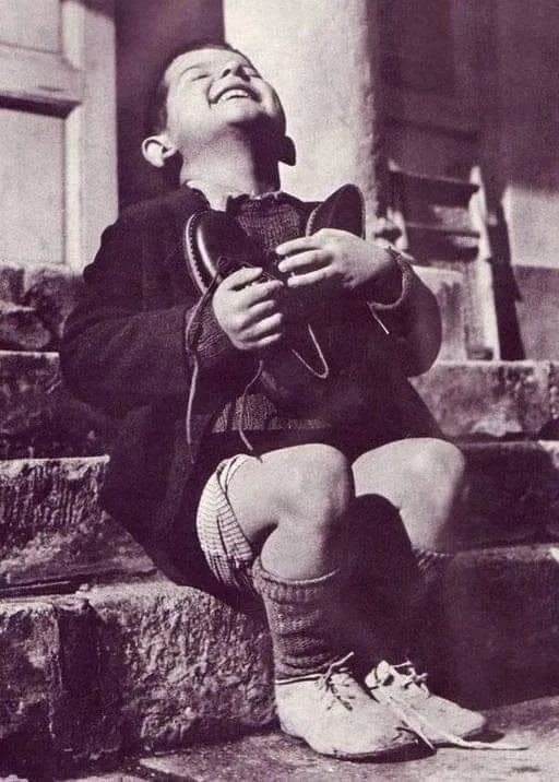 A vintage black-and-white photo shows a young boy sitting on stone steps, laughing joyfully with his head tilted back. He…