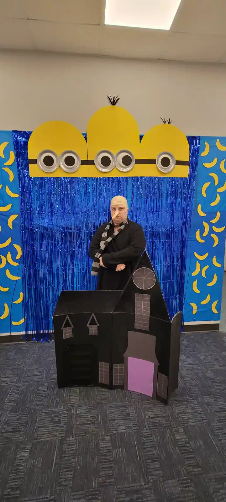 A person wearing a bald cap, glasses, and a long nose stands behind a black cardboard house with a purple door. Behind them…
