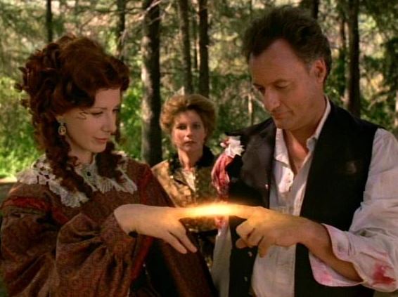 two people in period costumes hold hands over a glowing orb in a forest, while a third person watches. The man's shirt and…