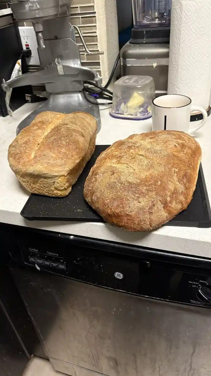 Two rustic, golden-brown loaves of homemade bread sit on a black cutting board atop a kitchen counter, next to a white mug…