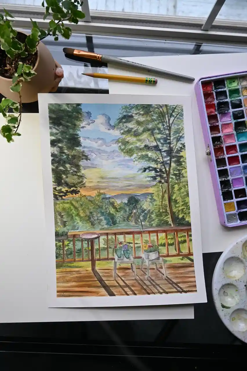 A watercolor painting of a wooden deck with chairs and a table, surrounded by trees and a distant landscape, sits on a desk…