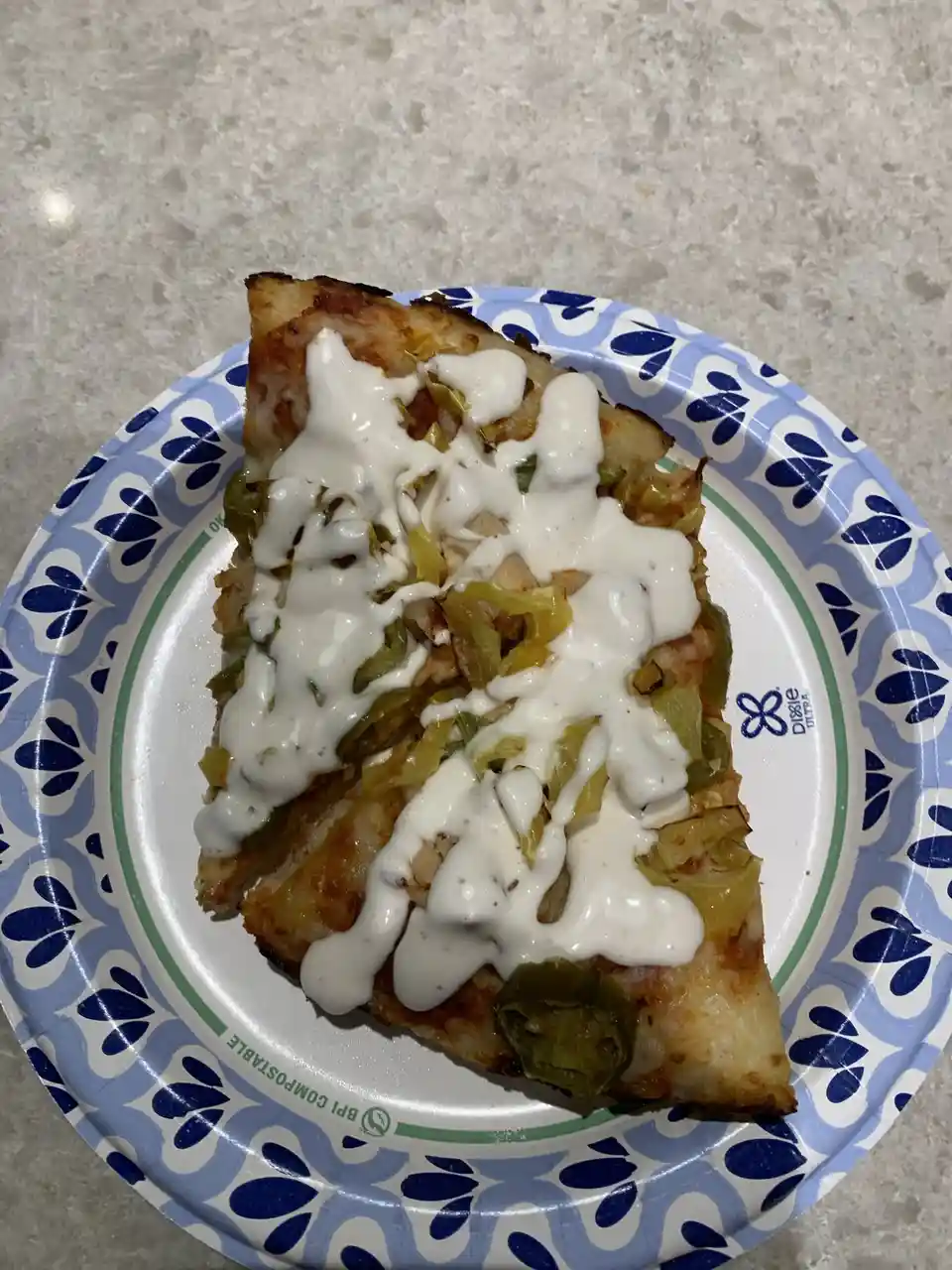 A single slice of pizza with a thick white sauce drizzled over it sits on a blue and white patterned Dixie Ultra paper…