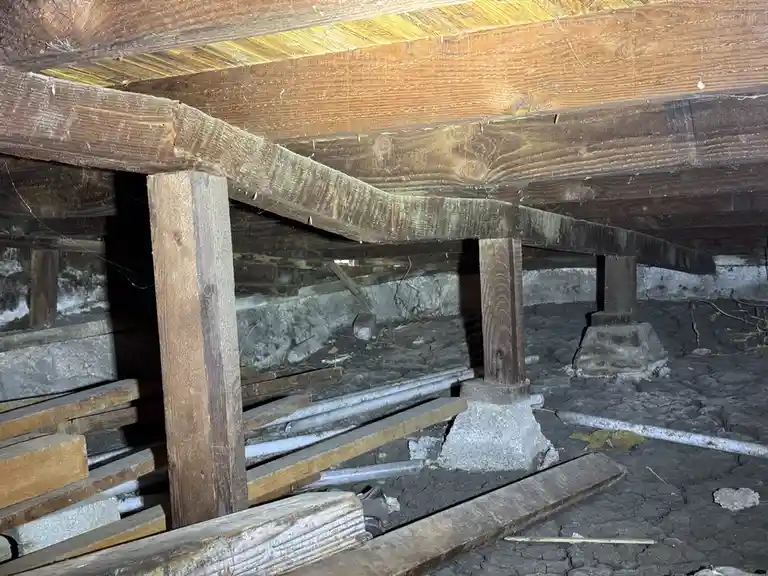 A dimly lit, dusty attic with exposed wooden beams and support posts, cracked dirt floor, and scattered debris. The overlaid…