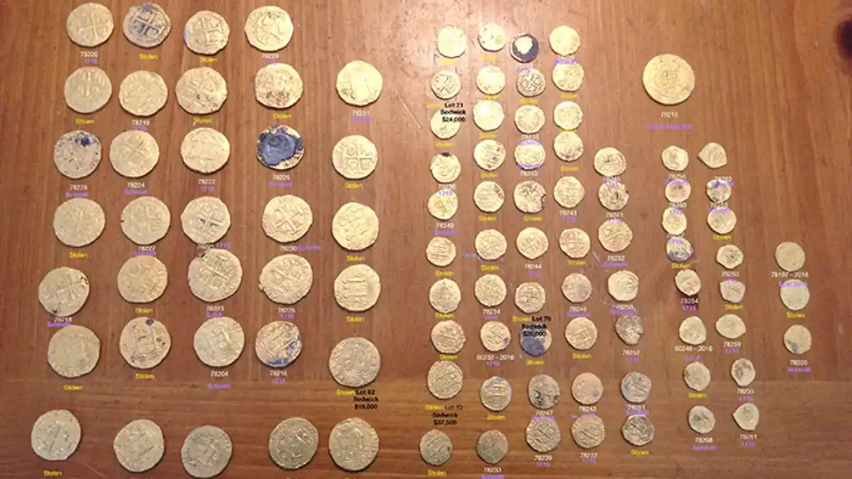 An overhead shot displays dozens of old, tarnished gold coins arranged on a wooden surface, each labeled with numbers…