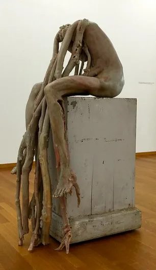 A sculpture of a humanoid figure with elongated, root-like limbs sits slumped atop a gray pedestal in a gallery. The…