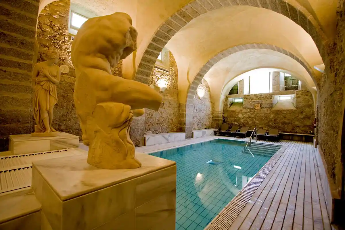 An indoor pool area with stone arches and classical statues, including a large, unfinished-looking male torso sculpture and…