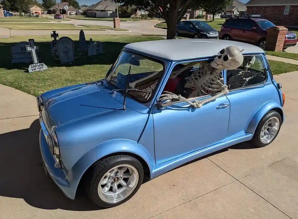 A light blue classic Mini Cooper is parked in a suburban driveway with two plastic skeletons seated inside, one leaning out…