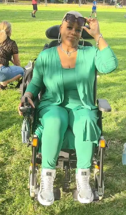 A woman in a green outfit and sunglasses sits confidently in a motorized wheelchair on a grassy field, adjusting her…