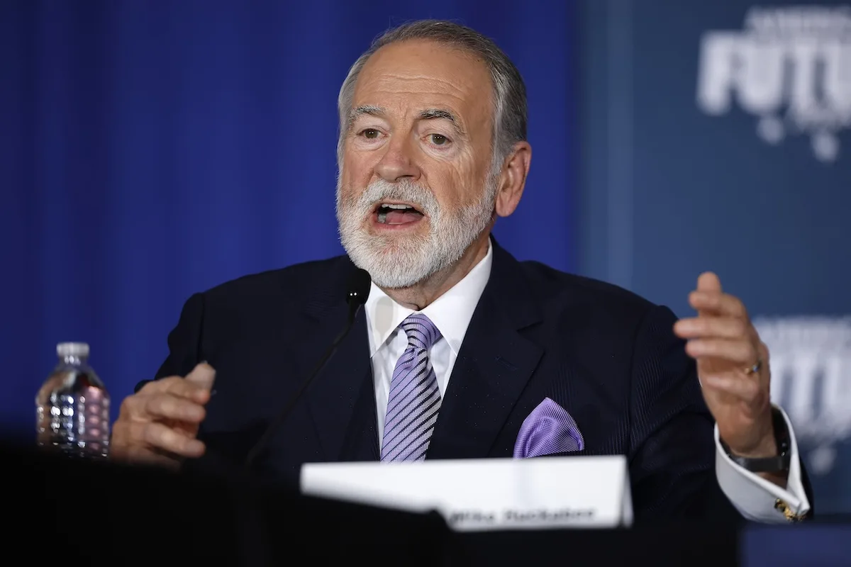 a man with a white beard wearing a dark suit and purple tie speaks at a podium with a microphone, gesturing with both hands…