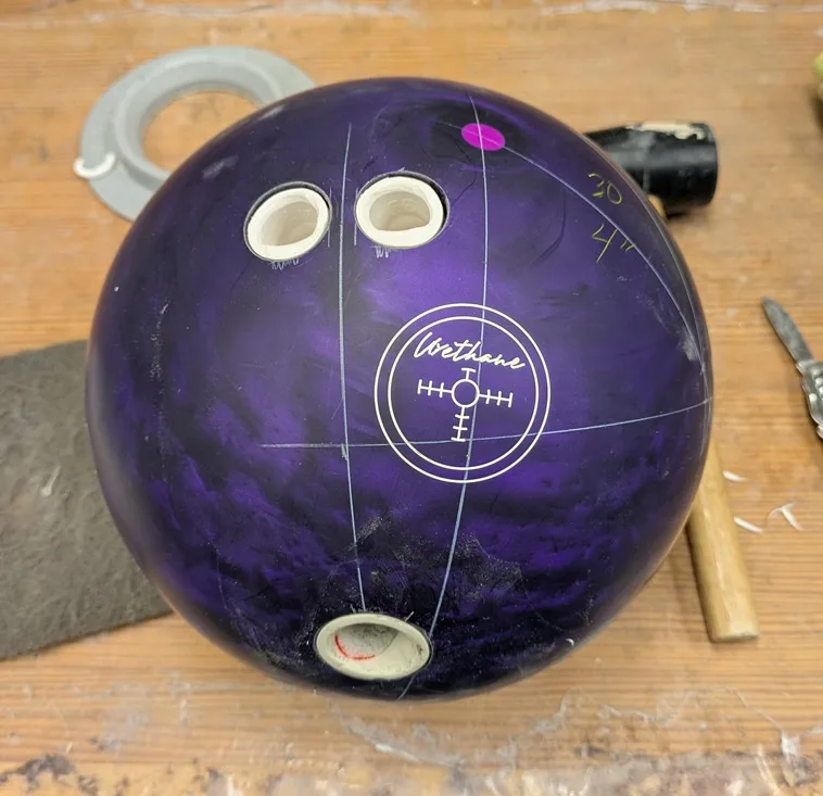 A purple bowling ball with three finger holes drilled into it sits on a wooden workbench, surrounded by tools like a knife…