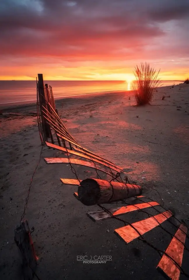 A serene beach sunset with vibrant orange and red skies over calm water, featuring a weathered wooden fence and log lying on…