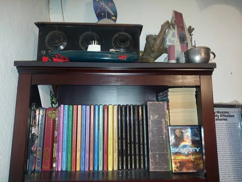 A wooden bookshelf filled with books, including a row of colorful spines, a stack of yellowed papers, and a game box. On top…