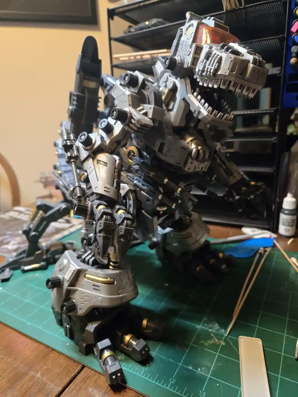 A highly detailed, silver and black robotic dinosaur model, resembling a T-Rex, is shown on a green cutting mat surrounded…
