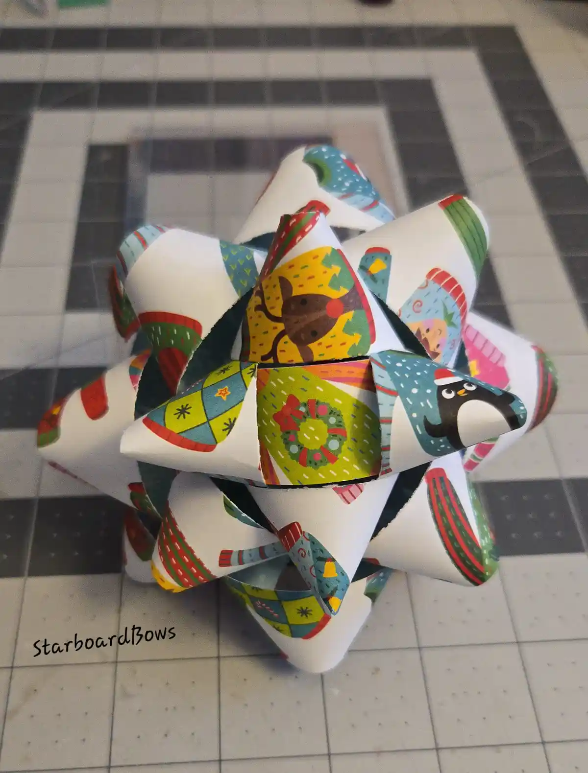 A colorful, multi-patterned Christmas bow made from paper with festive designs like reindeer, penguins, wreaths, and candy…