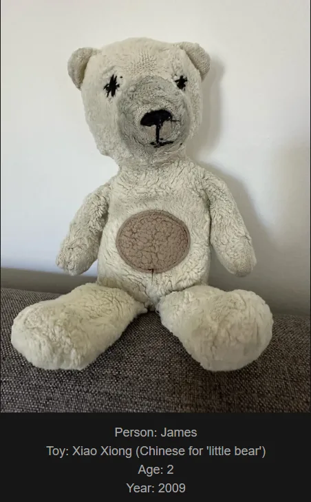 A worn, light-colored stuffed teddy bear with black marker eyes and nose sits on a textured gray surface. A circular patch…