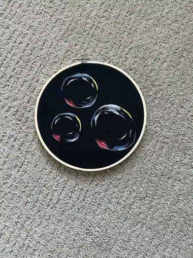 A circular embroidery hoop with black fabric holds an embroidered design of three stylized bubbles, each outlined in blue…