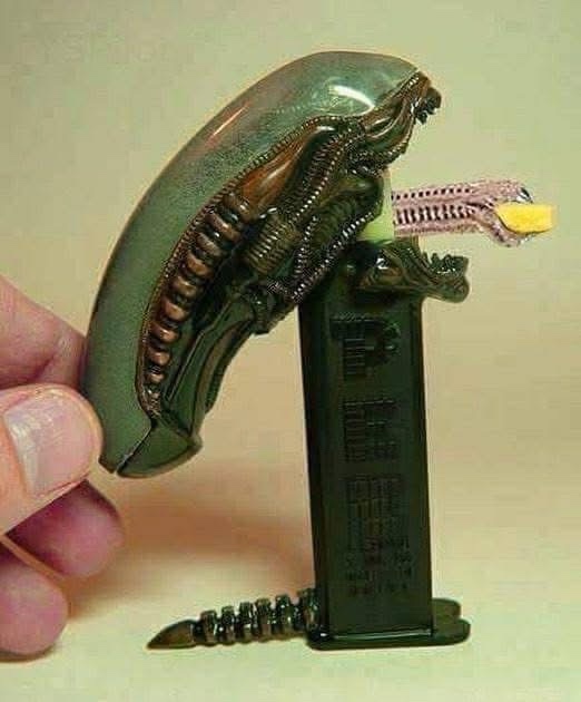 A hand holds a small, green, alien-like figurine with a zipper mouth, which is emerging from a black, rectangular object…