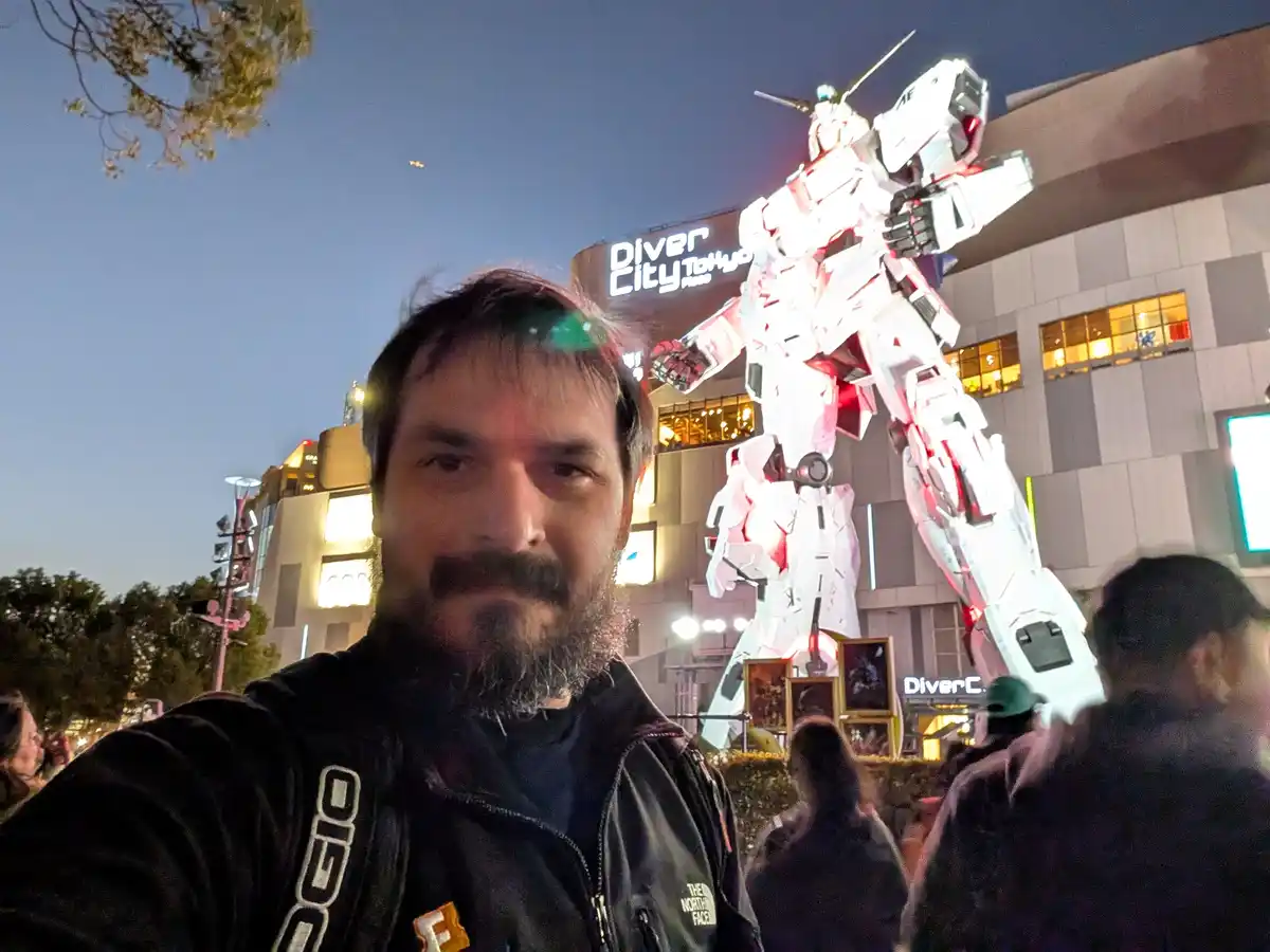 A man with a beard takes a selfie at night in front of a large, illuminated white and red Gundam statue outside Diver City…