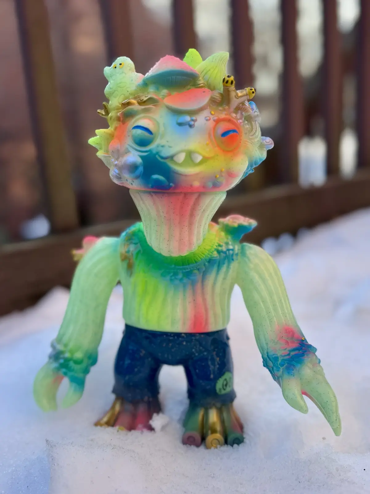 A colorful, translucent, rainbow-painted toy monster with multiple eyes, small creatures on its head, and clawed hands…
