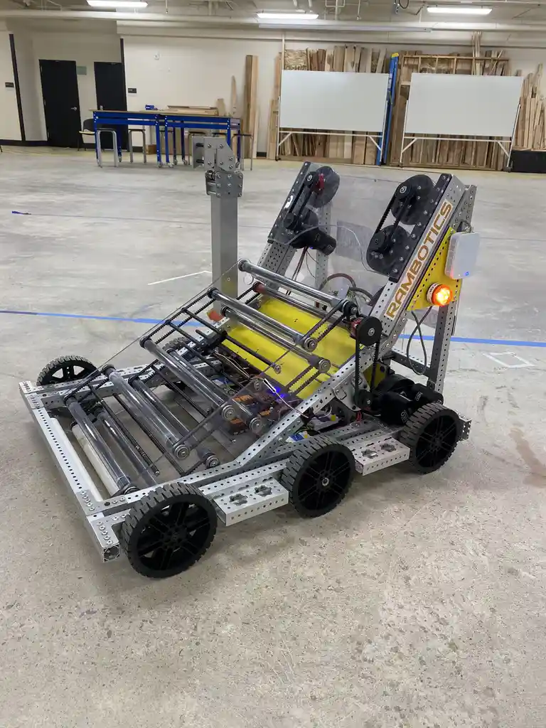 A custom-built robot with a silver metal frame, black wheels, and a yellow central component, sits on a concrete floor in a…