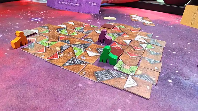 A tabletop view of the board game 'Viticulture: Manufacturing' with colorful wooden player pieces (orange, green, purple)…