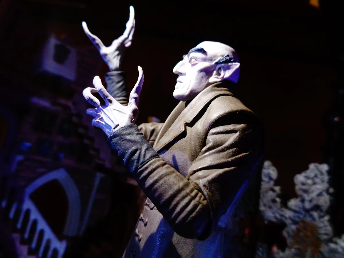 A statue of a bald, pale-skinned vampire with pointed ears and long fingernails, wearing a brown coat and black gloves…