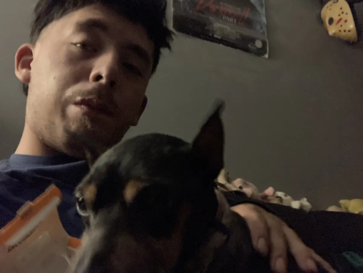 A man with dark hair and a goatee, wearing a blue shirt, is seen resting his head near a small black and tan dog, possibly a…