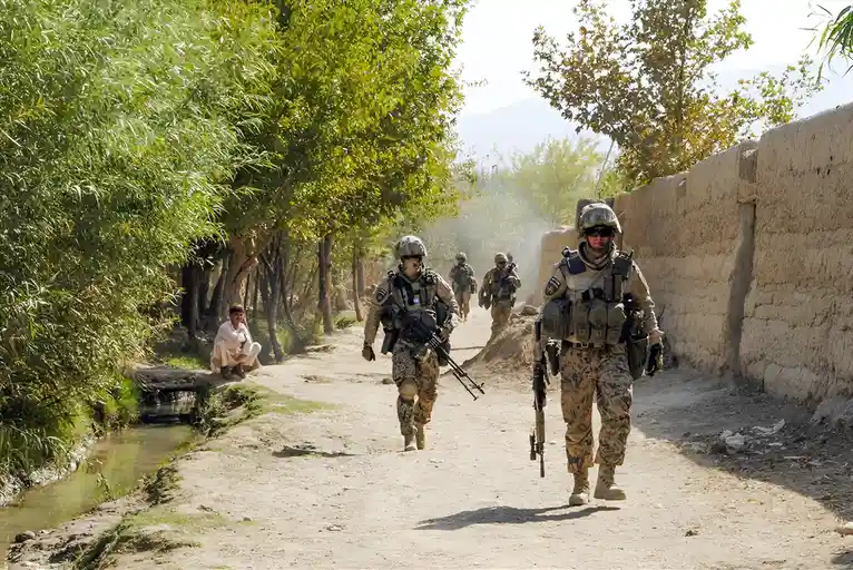A group of heavily armed soldiers in camouflage gear march down a dusty rural path, kicking up dust. On the left, a man in…