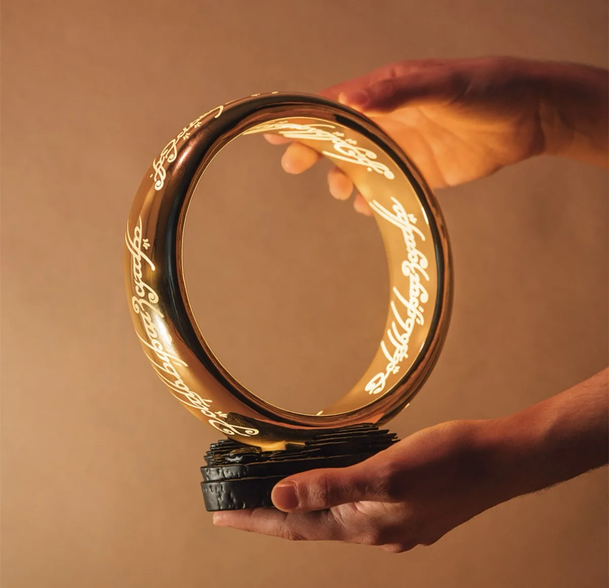 A person holds a large, glowing golden ring with Elvish script engraved around its circumference, resting on a black…
