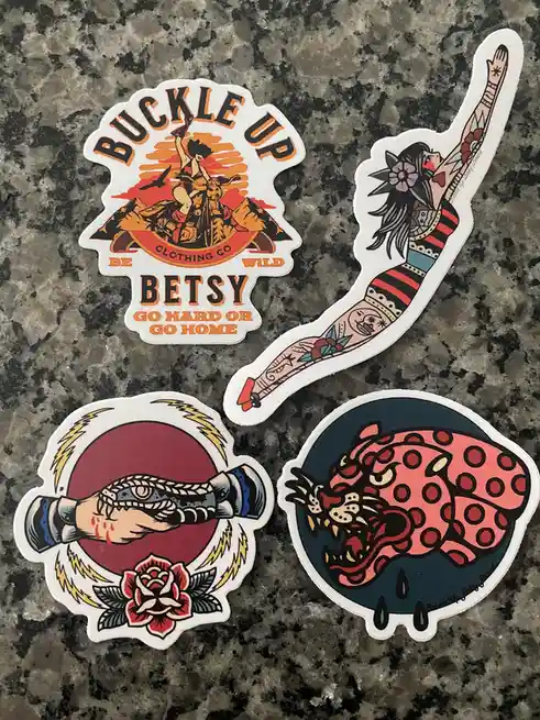 Four stickers from 'Buckle Up Betsy' clothing brand lie on a granite countertop. One shows a biker girl with 'Buckle Up…