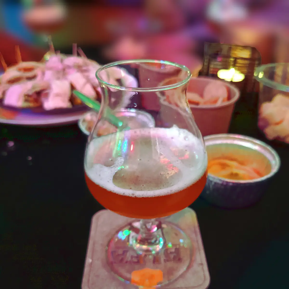 A close-up photo of a beer glass with a foamy head, sitting on a coaster on a dark table in a dimly lit, colorful bar…