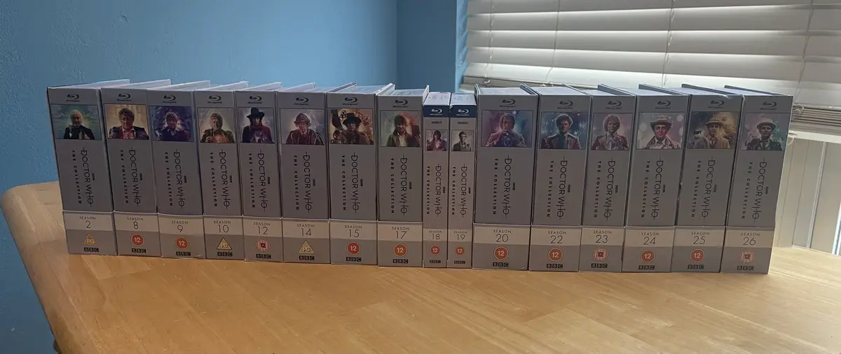 A row of Doctor Who Blu-ray box sets, labeled 'The Collection', are arranged on a wooden table. The spines show seasons 2…