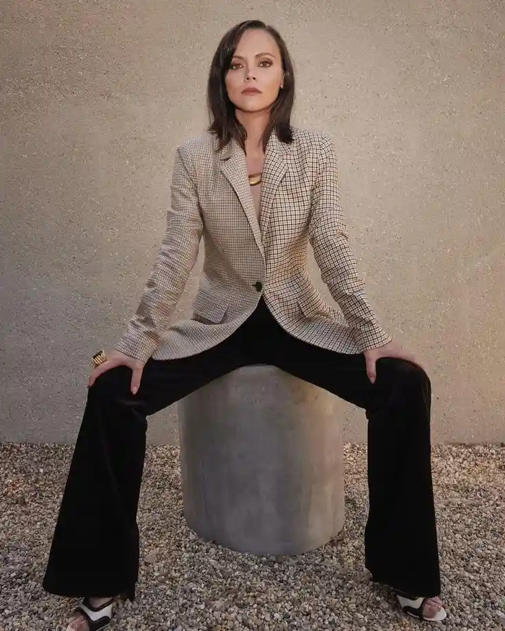 A woman with dark hair sits on a cylindrical concrete block outdoors, wearing a beige and black checkered blazer, black…