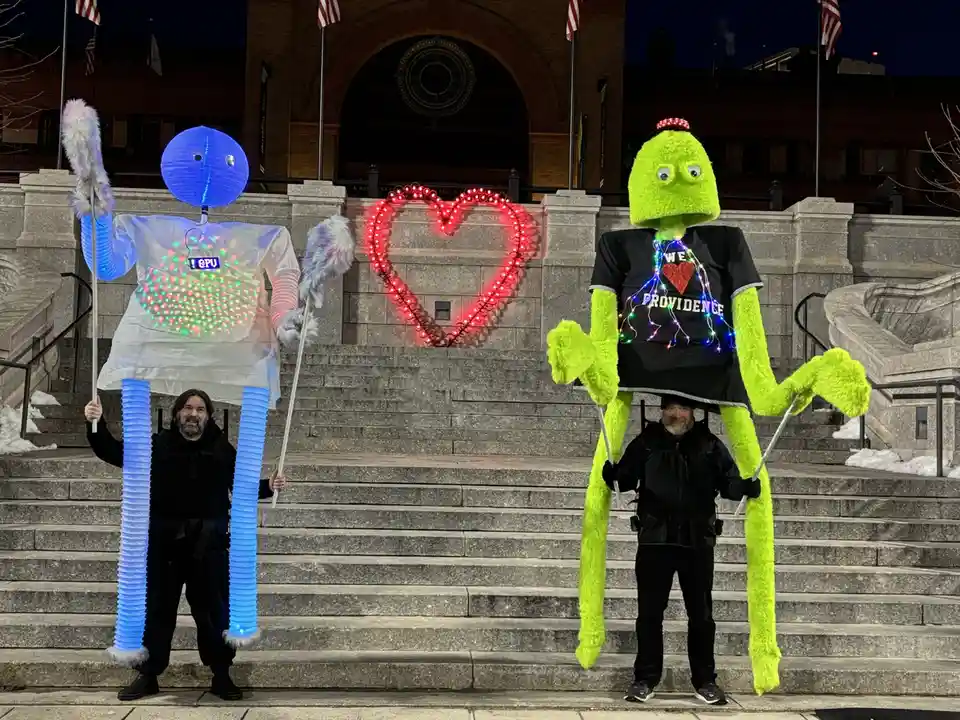 Two people in giant costumes stand on stone steps in front of a building with a red heart light display. The left costume is…