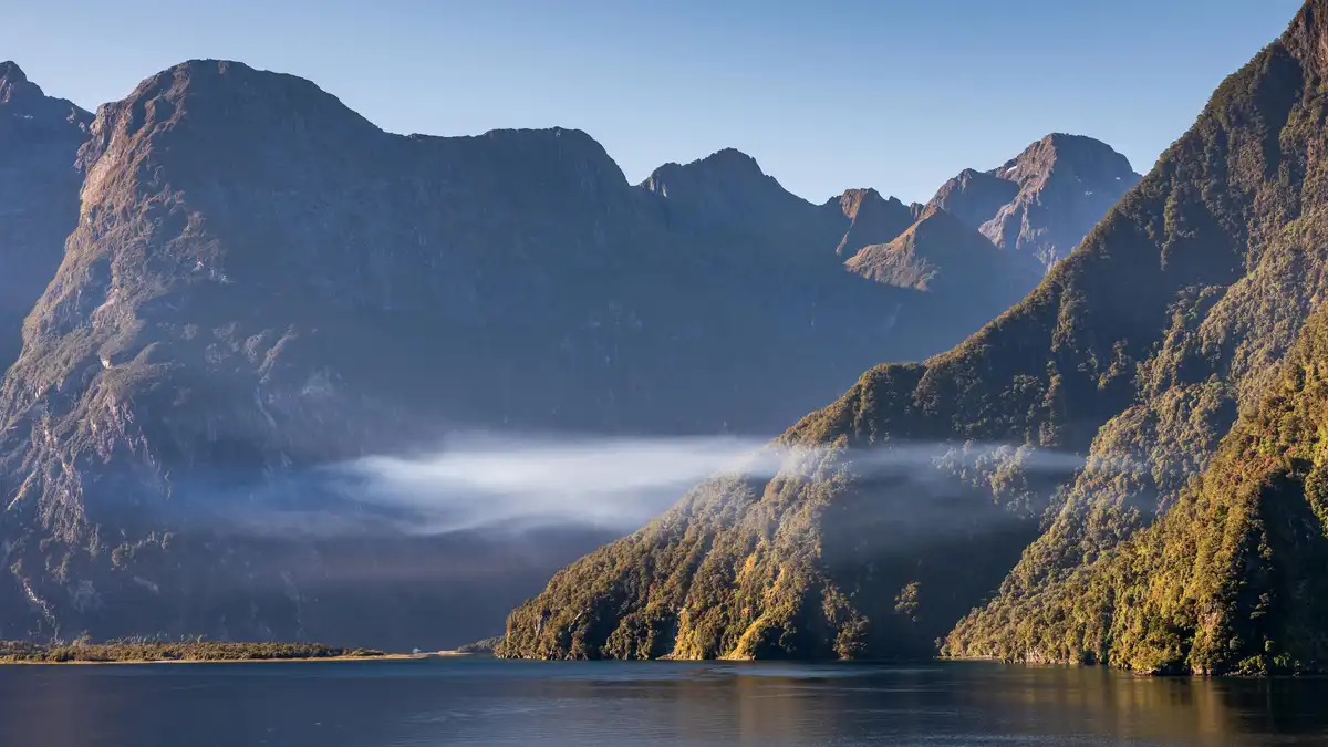 Fiery sunrise casts a warm glow on misty Fiordland mountains, shrouding the tranquil lake in an ethereal veil…