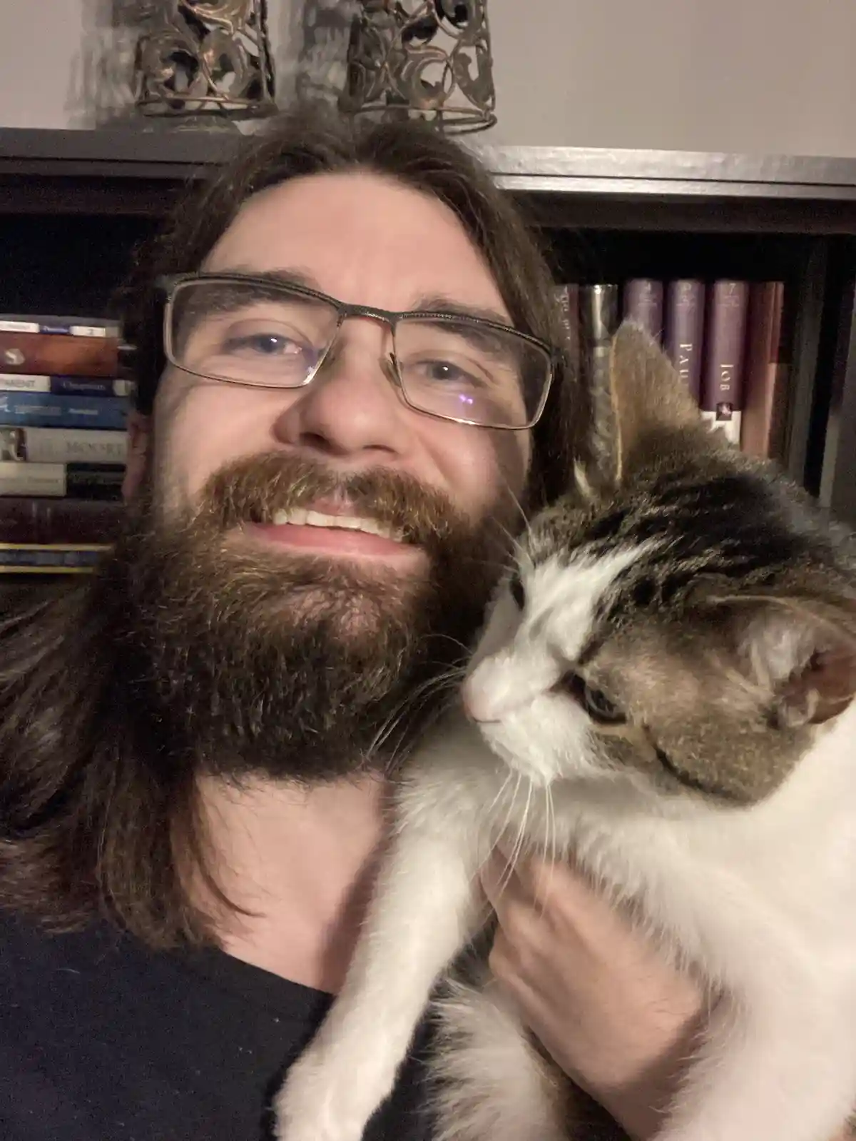 A man with long dark hair, glasses, and a beard smiles at the camera while holding a white and gray tabby cat. The cat looks…