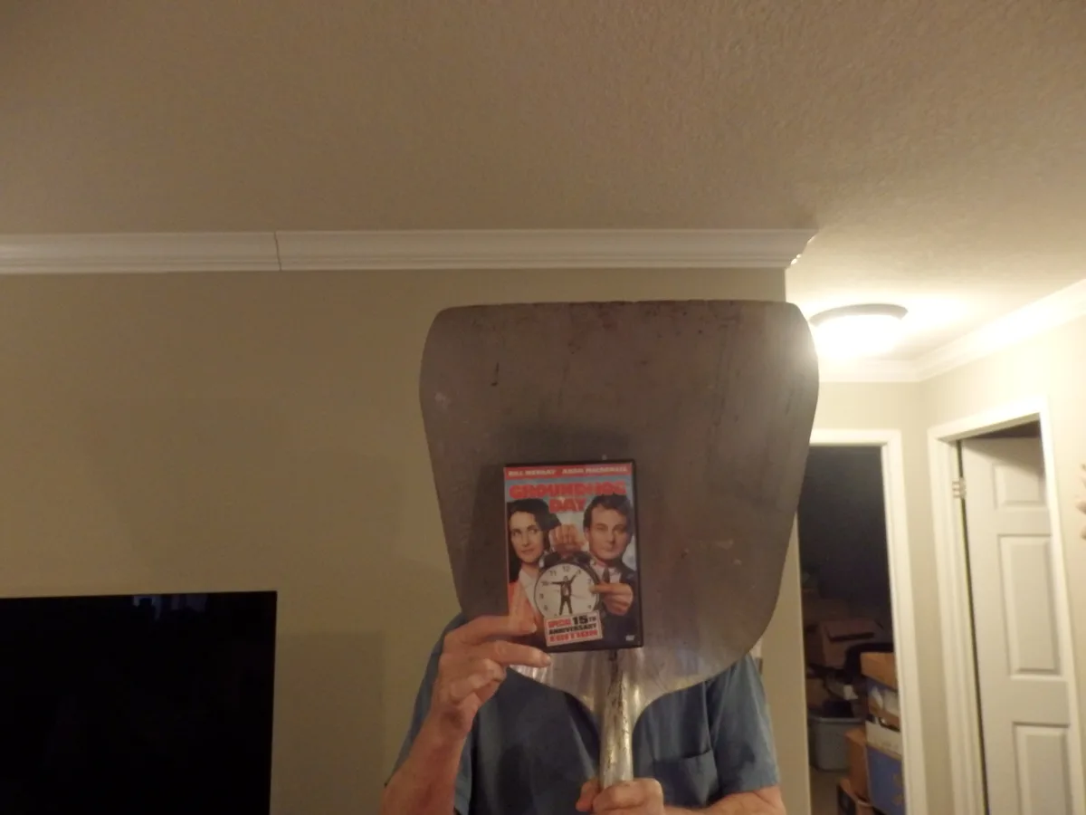 A person holds a large, dirty shovel in front of their face, obscuring it completely, while holding up a DVD case for…