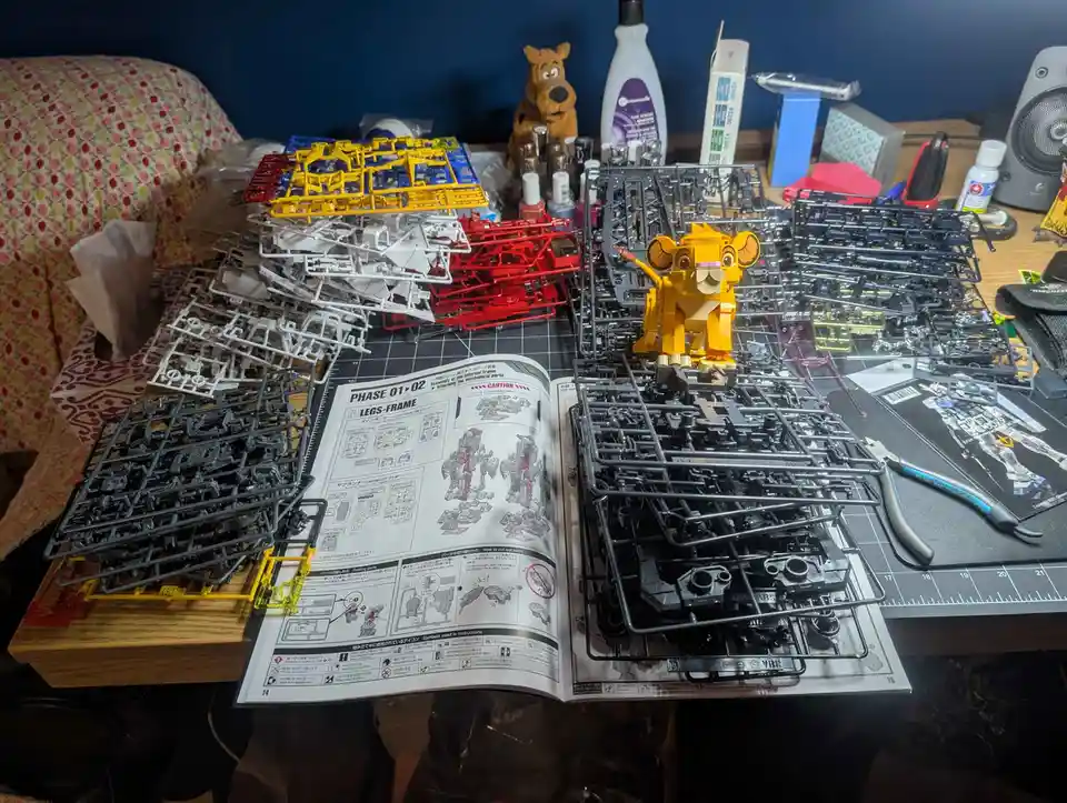 A cluttered desk is covered with numerous unassembled plastic model kits in various colors, including stacks of gray, white…