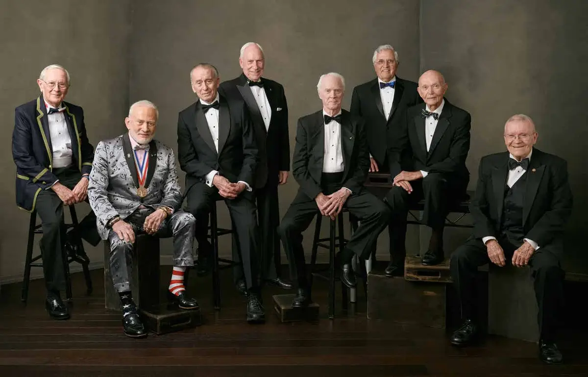 A formal group portrait of eight elderly men in tuxedos posing on stools and crates. One man in the center wears a flashy…