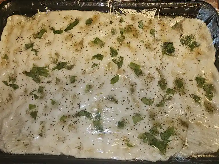 A close-up of raw pizza dough in a black baking tray, covered with plastic wrap, showing visible bubbles, scattered green…