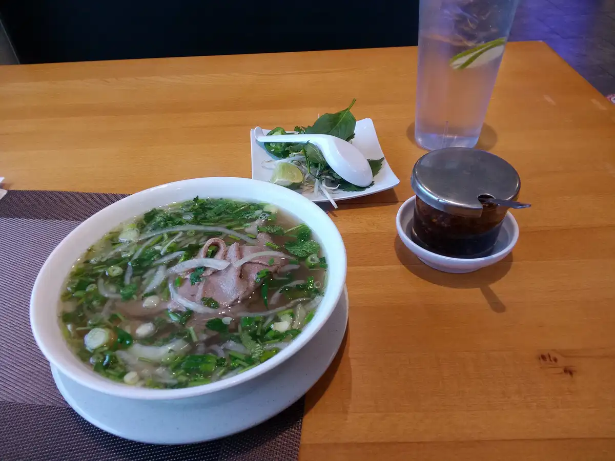 A bowl of Vietnamese pho with rice noodles, thinly sliced beef, green herbs, and onions sits on a wooden table, accompanied…