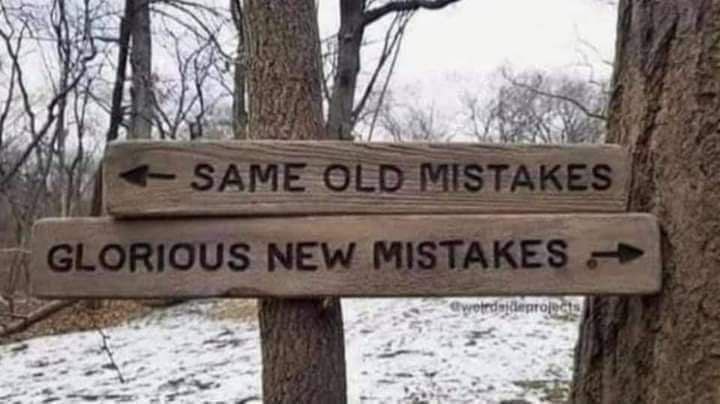 Two wooden signs nailed to tree trunks in a snowy, wooded area. The top sign points left and reads 'SAME OLD MISTAKES'. The…